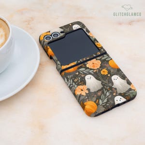 Könnte beinhalten: Eine Handyhülle mit einem dunklen Hintergrund und einem Halloween-Muster. Das Design umfasst orangefarbene Kürbisse, Blumen und weiße Geisterillustrationen. Die Hülle ist geöffnet und zeigt einen schwarzen Bildschirm. Der Markenname "GLITCHGLAMCO" ist sichtbar.