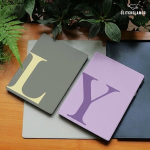 Pode incluir: Capas para laptop em cinza, lavanda e preto, cada uma com uma letra grande. As letras são em cores contrastantes. As capas estão dispostas sobre uma superfície de madeira com folhagem. As capas são projetadas para um laptop.