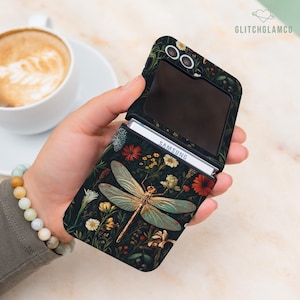 Może przedstawiać: Czarne etui na telefon z motywem ważki i kwiatów. Etui jest otwarte, ukazując telefon Samsung. Ważka ma złote i turkusowe skrzydła. Etui jest ozdobione czerwonymi, żółtymi i białymi kwiatami.