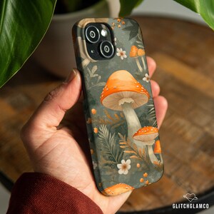 May include: A phone case featuring a whimsical mushroom design. The case has a dark green background with orange and white mushrooms, white flowers, and fern-like leaves. The case is held in a person's hand.