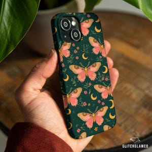 May include: A dark green phone case featuring a repeating pattern of pink moths, yellow crescent moons, and small pink flowers. The case has a glossy finish and is held in a hand. The brand name "GLITCHGLAMCO" is visible in the bottom right corner.