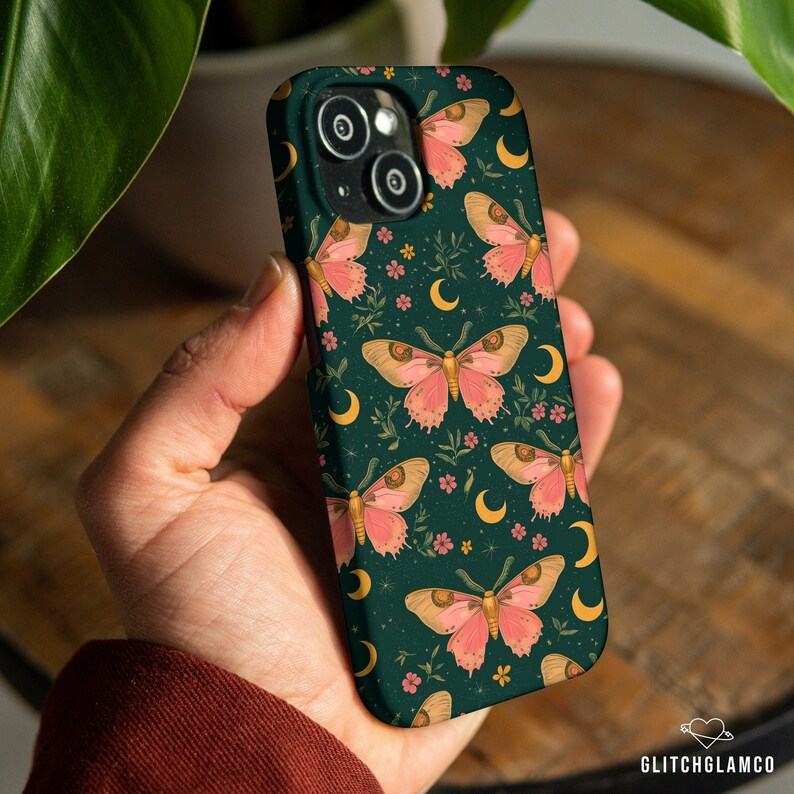 May include: A dark green phone case featuring a repeating pattern of pink moths, yellow crescent moons, and small pink flowers. The case has a glossy finish and is held in a hand. The brand name "GLITCHGLAMCO" is visible in the bottom right corner.