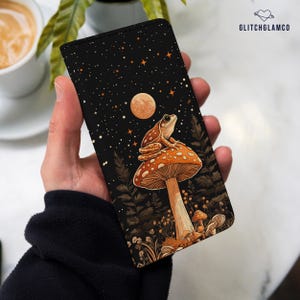 Peut inclure: Une coque de téléphone noire avec une illustration fantaisiste d'une grenouille assise sur un champignon sous une pleine lune et des étoiles. Le champignon est orange et brun avec des points blancs. La grenouille est verte avec des points bruns. L'arrière-plan est bleu foncé avec des étoiles blanches.