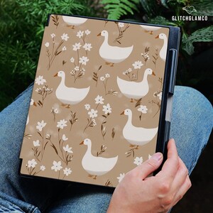 May include: A black tablet case featuring a repeating pattern of white geese and delicate white flowers on a tan background. The case is held in a person's lap, with the text "GLITCHGLAMCO" visible in the upper right corner.