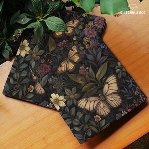 May include: Two rectangular decorative mats with a dark background featuring a floral and butterfly design. The butterflies are cream and brown, and the flowers are yellow, purple, and burgundy. The mats are on a wooden surface.