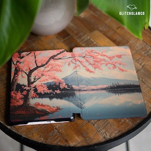 以下が含まれることがあります： ピンクの桜の花が咲く山の風景が描かれた、白黒の電子書籍リーダーカバー。カバーが開いて、電子書籍リーダーの画面が見えるようになっています。