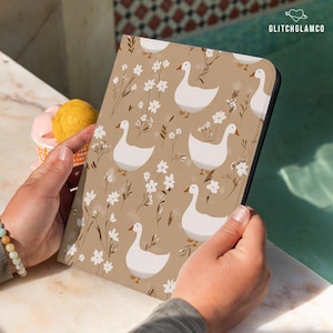 Puede incluir: Una funda para tableta de color beige con un estampado de gansos blancos y flores blancas. Los gansos tienen picos y patas naranjas. Las flores y el follaje son en tonos marrones y blancos. La funda la sostiene una persona.