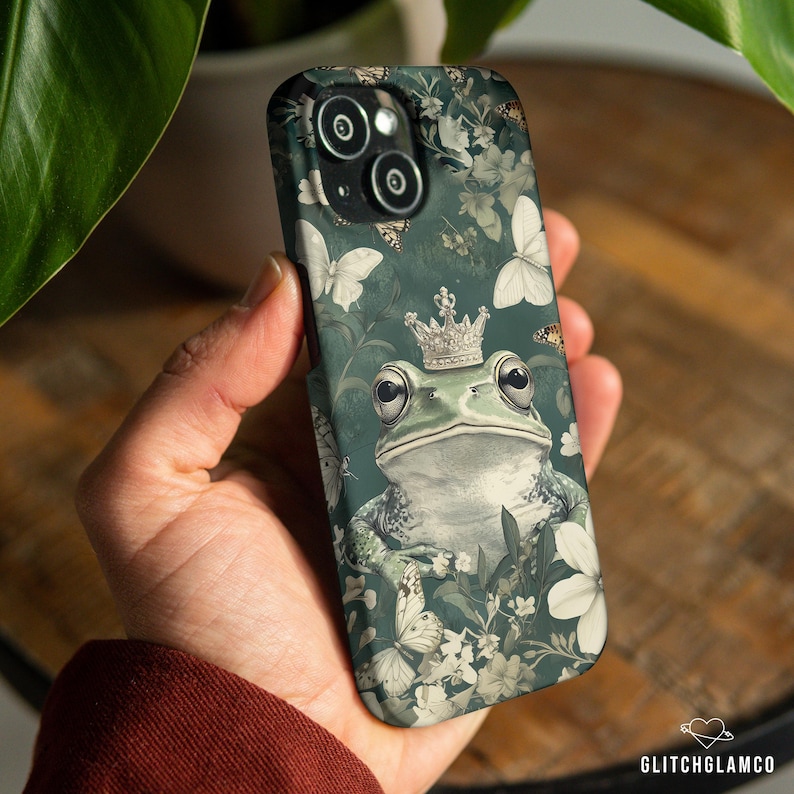May include: A dark green phone case featuring a frog wearing a crown, surrounded by butterflies and flowers. The case has a matte finish and a whimsical design. The phone case is held in a hand.