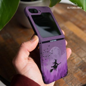 Puede incluir: Funda morada para teléfono Samsung con diseño de Halloween. La funda presenta la silueta de una bruja en una escoba volando frente a una luna llena, con un bosque de fondo. El teléfono está abierto, mostrando la pantalla.