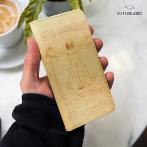 May include: A rectangular beige phone case featuring a printed illustration of the Vitruvian Man by Leonardo da Vinci, complete with the original mirror-writing text surrounding the figure. The brand name GLITCHGLAMCO is printed in the top right corner.