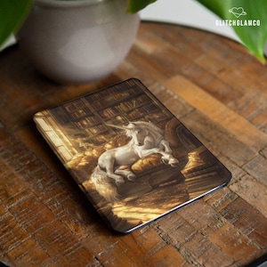 Peut inclure: Un étui rectangulaire avec une illustration détaillée d'une licorne blanche se reposant sur des livres dans une bibliothèque. L'image a des tons chauds et dorés et un thème fantastique. L'étui est sur une surface en bois.