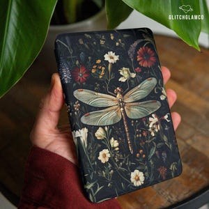 May include: A black phone case featuring a detailed illustration of a dragonfly with delicate wings, set against a backdrop of colorful flowers and foliage. The case has the text "GLITCHGLAMCO" in the upper right corner.