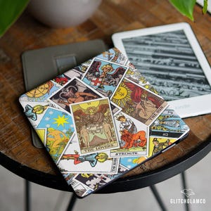 Op de afbeelding: Een Kindle e-reader met een kleurrijke hoes met tarotkaartontwerpen, waaronder "The Lovers" en "Strength". De hoes ligt op een houten tafel naast een grijze hoes en een andere Kindle. Op de achtergrond is een plant te zien.