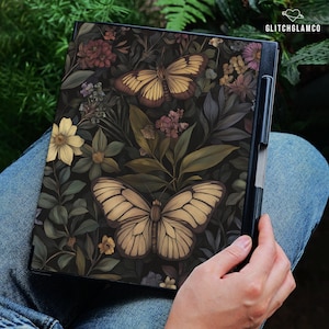 May include: A black notebook featuring a detailed illustration of two butterflies and various flowers. The butterflies have tan and brown wings, set against a dark background of leaves and blooms. The notebook is held in a person's lap.