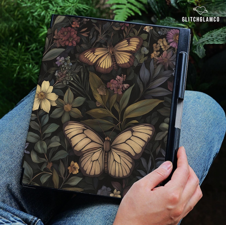 May include: A black notebook featuring a detailed illustration of two butterflies and various flowers. The butterflies have tan and brown wings, set against a dark background of leaves and blooms. The notebook is held in a person's lap.