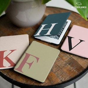 Könnte beinhalten: Eine Sammlung von Kindle E-Reader-Hüllen in verschiedenen Farben, jede mit einem großen, fetten Buchstaben. Die Hüllen sind in Grün-, Rosa- und Beigetönen gehalten, mit weißen, roten und schwarzen Buchstabendesigns. Eine Hülle ist geöffnet und zeigt den E-Reader-Bildschirm.