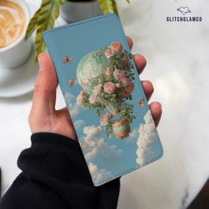 May include: A rectangular phone case featuring a light blue sky background with white clouds and a hot air balloon adorned with pink roses and green foliage. Small orange butterflies are scattered around the balloon. The text GLITCHGLAMCO is visible.