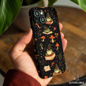 May include: Black phone case featuring a repeating pattern of cartoon frogs wearing witch hats and sitting on books. The design includes orange mushrooms, stars, and leaves. The case has a glossy finish and is held in a hand.