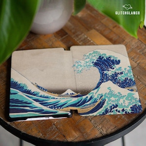 Könnte beinhalten: Eine Handyhülle mit dem Kunstwerk "Die große Welle vor Kanagawa". Das Design zeigt eine große blaue Welle mit weißen Schaumkronen, Booten und einem Berg im Hintergrund. Die Hülle ist beige und trägt den Text "GLITCHGLAMCO".