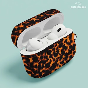 Może przedstawiać: Otwarty futerał na AirPods z wzorem w kolorze skorupy żółwia w odcieniach brązu i pomarańczy. Futerał mieści dwa białe AirPods. Nazwa marki "GLITCHGLAMCO" jest wydrukowana na białym tle w prawym górnym rogu obrazu.