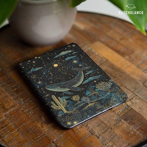Può includere: Custodia rettangolare nera con un design di balena celeste. La custodia presenta una balena che nuota in un cielo stellato con cactus e piante del deserto. Il design include accenti oro e verde acqua. Il marchio "GLITCHGLAMCO" è visibile.
