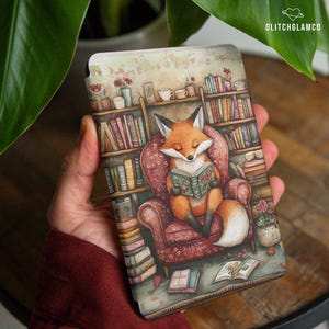May include: A decorative phone case featuring a watercolor illustration of a fox reading a book in a cozy armchair. The background includes a bookshelf, flowers, and open books, creating a whimsical, literary theme. The case has the text "GLITCHGLAMCO".