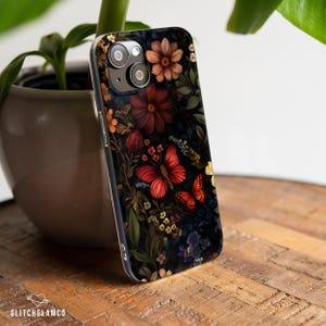 May include: A smartphone case with a dark floral pattern, including red butterflies and flowers. The case has a black backdrop with various colourful flowers and foliage. The brand name "GLITCHGLAMCO" is visible at the bottom.
