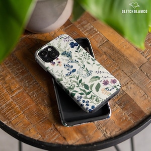 May include: A white phone case with a floral pattern featuring green leaves, blue and purple flowers, and white accents. The case is designed for a phone with a triple camera lens.