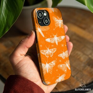 May include: Orange phone case featuring a white moth and dragonfly pattern. The case has a glossy finish and a protective design. The phone case is held in a person's hand. The phone case has a camera cutout.