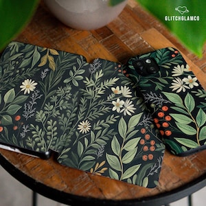 May include: A collection of phone cases and a notebook featuring a dark background with a botanical pattern. The design includes white daisies, green leaves, and orange berries. The cases and notebook are arranged on a wooden surface.