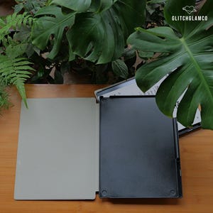 May include: An open black tablet case with a light gray interior sits on a wooden surface. The case is next to a tablet with a white screen. Lush green plants are in the background. The text "GLITCHGLAMCO" is in the upper right corner.