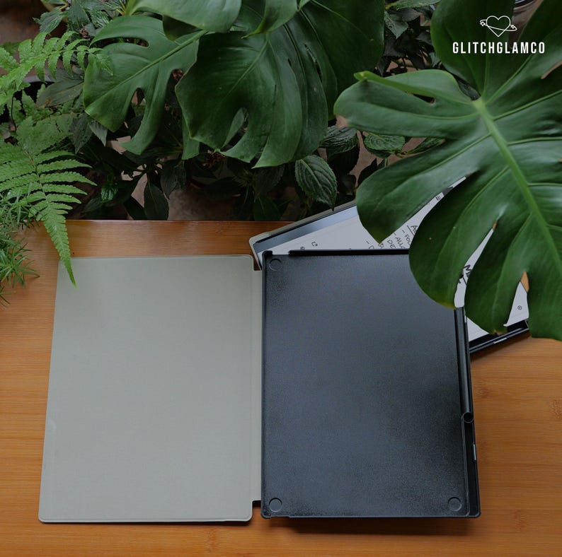 May include: An open black tablet case with a light gray interior sits on a wooden surface. The case is next to a tablet with a white screen. Lush green plants are in the background. The text "GLITCHGLAMCO" is in the upper right corner.