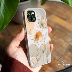 Puede incluir: Una funda de teléfono blanca con un diseño floral. Las flores están delineadas en negro y tienen acentos naranja y beige. La funda tiene un recorte para la cámara y una parte posterior transparente.