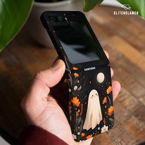 Pode incluir: Capa de telefone Samsung preta com um design de fantasma e floral. A capa apresenta um fundo preto com flores laranja, flores brancas e uma lua cheia. O telefone está aberto, revelando a tela.