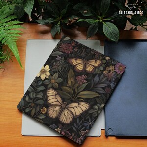May include: A decorative notebook with a dark floral and butterfly design. The cover features a pattern of flowers and butterflies in shades of cream, brown, and green. The notebook is resting on a light gray surface.