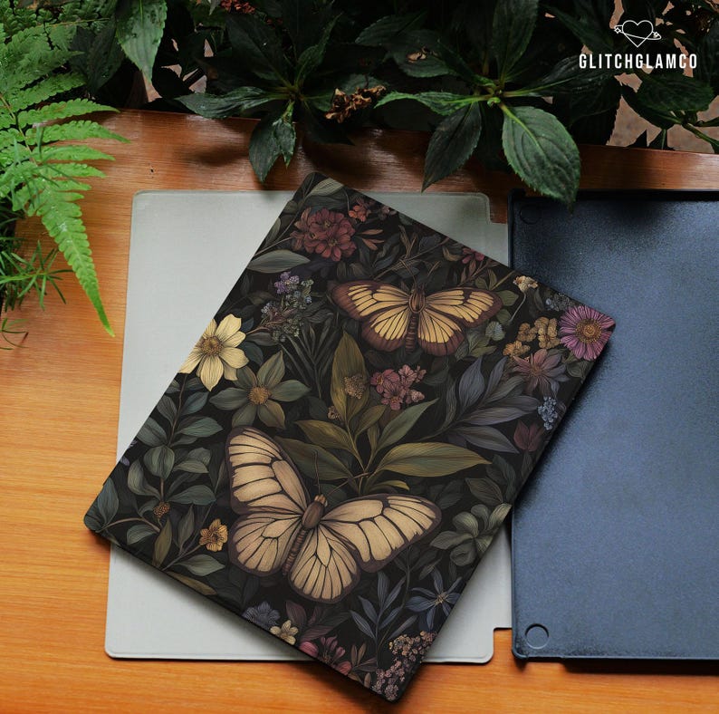 May include: A decorative notebook with a dark floral and butterfly design. The cover features a pattern of flowers and butterflies in shades of cream, brown, and green. The notebook is resting on a light gray surface.