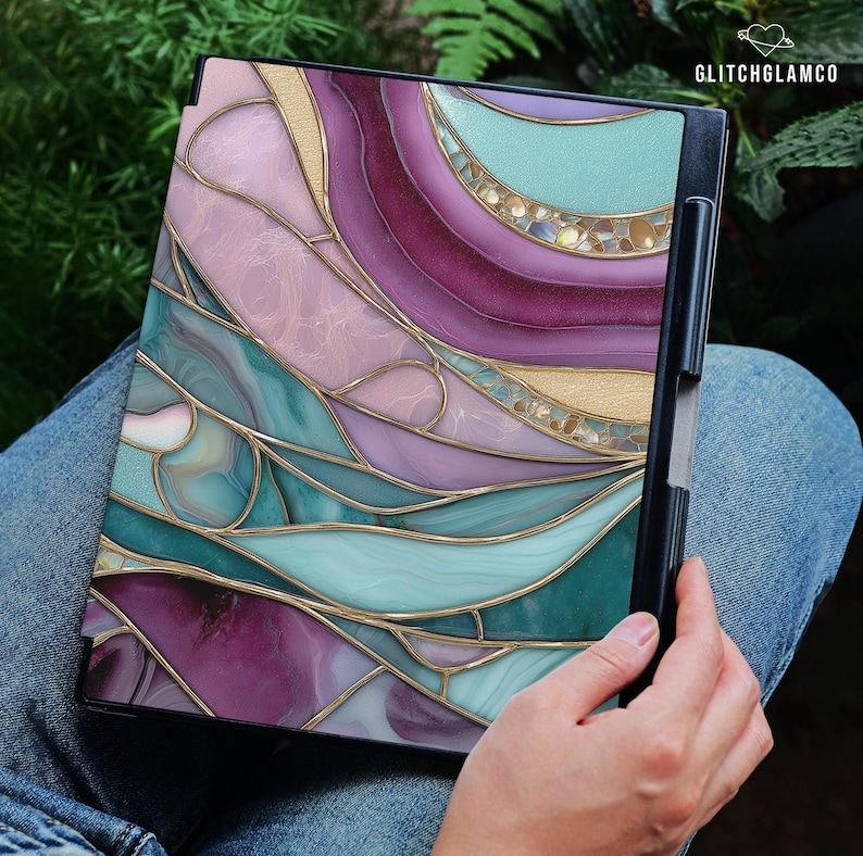 May include: A laptop case featuring a stained glass design with swirling patterns in shades of purple, teal, and gold. The case has a black frame and is held by a person wearing blue jeans. The text "GLITCHGLAMCO" is visible in the upper right corner.