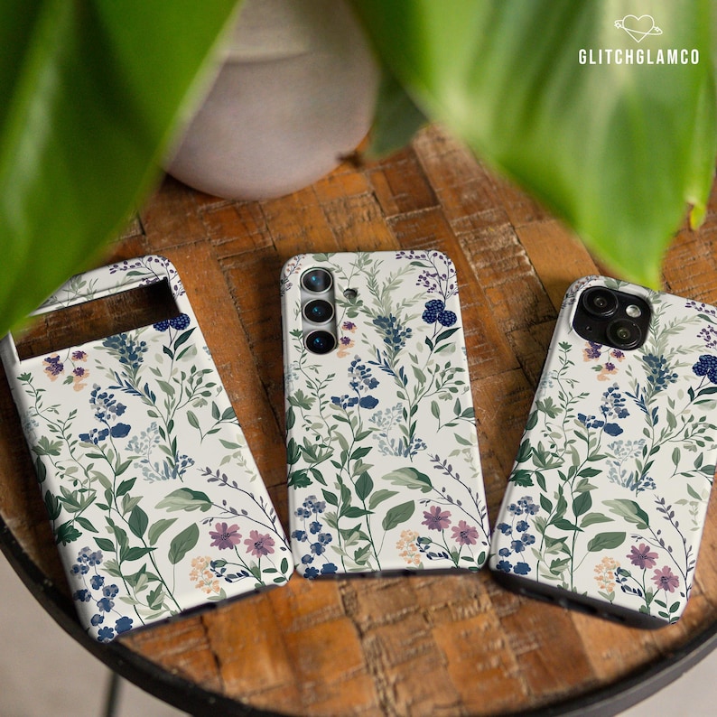 May include: Three phone cases with a white background and a floral pattern in shades of blue, green, and purple. The cases have a variety of floral designs, including small flowers, leaves, and berries.