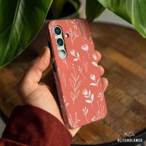 May include: Coral-colored phone case featuring a white floral pattern. The design includes various stylized flowers and leaves. The case has a camera module with three lenses. The GlitchGlamCo logo is visible in the bottom right corner.