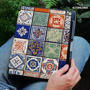 Può includere: Una custodia nera per tablet con un design a mosaico. Le piastrelle sono quadrate e presentano vari motivi nei toni del blu, arancione, verde e bianco. La custodia è tenuta da una persona che indossa jeans blu. Il testo "GLITCHGLAMCO" è visibile.