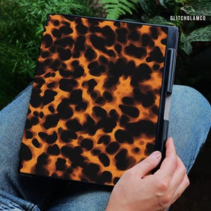 May include: A notebook with a tortoiseshell pattern cover in shades of brown and black. The notebook is held by a person wearing blue jeans. The text "GLITCHGLAMCO" is visible in the upper right corner.