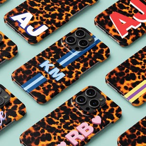 May include: A collection of personalized phone cases with a leopard print design. Each case features initials in various colors, including white, red, and pink. Some cases have stripes in blue, yellow, and purple.