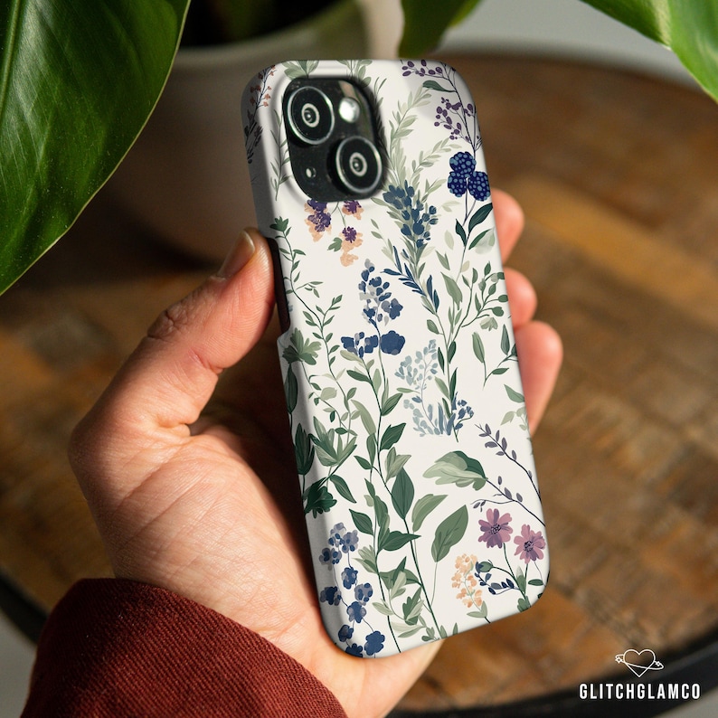 May include: A white phone case with a floral pattern featuring green leaves, blue and purple flowers, and white background.