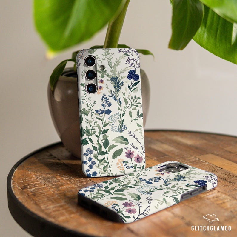 May include: Two phone cases with a floral pattern. The cases are white with green leaves, blue flowers, and purple berries. The case on the left has a camera cutout.