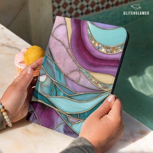 May include: A tablet case with a stained glass design. The case features a vibrant abstract pattern in shades of purple, teal, and gold. The design is reminiscent of a mosaic or a gemstone, with flowing lines and color gradients. The text "GLITCHGLAMCO" is visible.