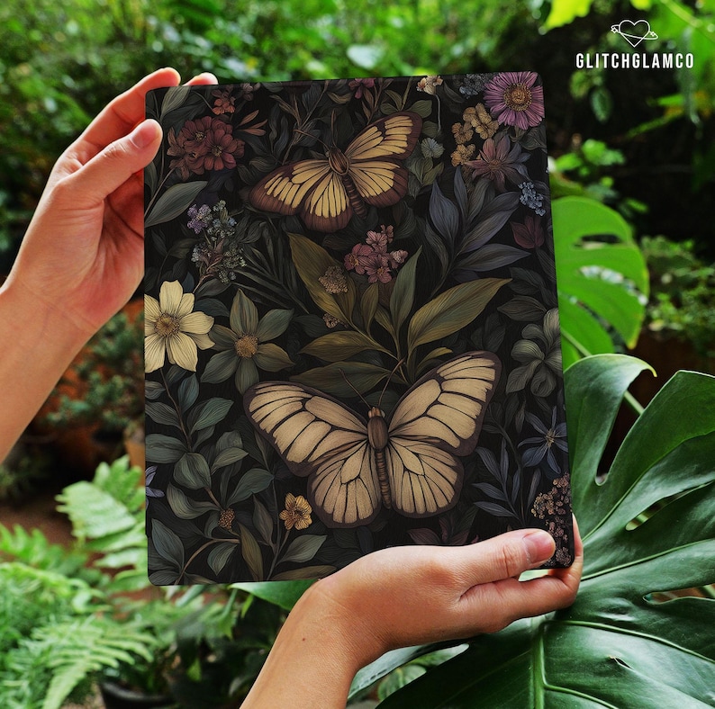 May include: A rectangular art print featuring a dark background with a botanical design. Two large butterflies with cream-colored wings are prominent, surrounded by flowers and foliage in shades of green, purple, and yellow. The print is held by hands.