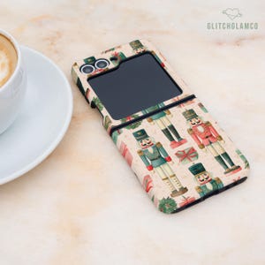 May include: A phone case with a festive Christmas theme. The case features a repeating pattern of toy nutcracker soldiers, wrapped gifts, and greenery on a beige background. The phone case is open, revealing the phone's screen.