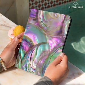 May include: A tablet case featuring an iridescent abalone shell design in shades of purple, green, and pink. The case has a black border and the text "GLITCHGLAMCO" in the upper right corner. The case is held by a person.