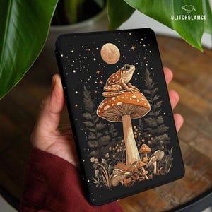 May include: Black phone case featuring a whimsical illustration of a frog perched atop a mushroom under a full moon and starry night sky. The design includes detailed forest elements in shades of brown and beige.