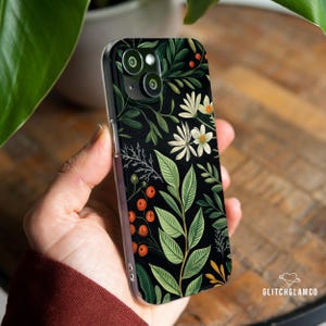 May include: A clear phone case featuring a black background with a floral design. The design includes white flowers, green leaves, and orange berries. The case is held in a person's hand.